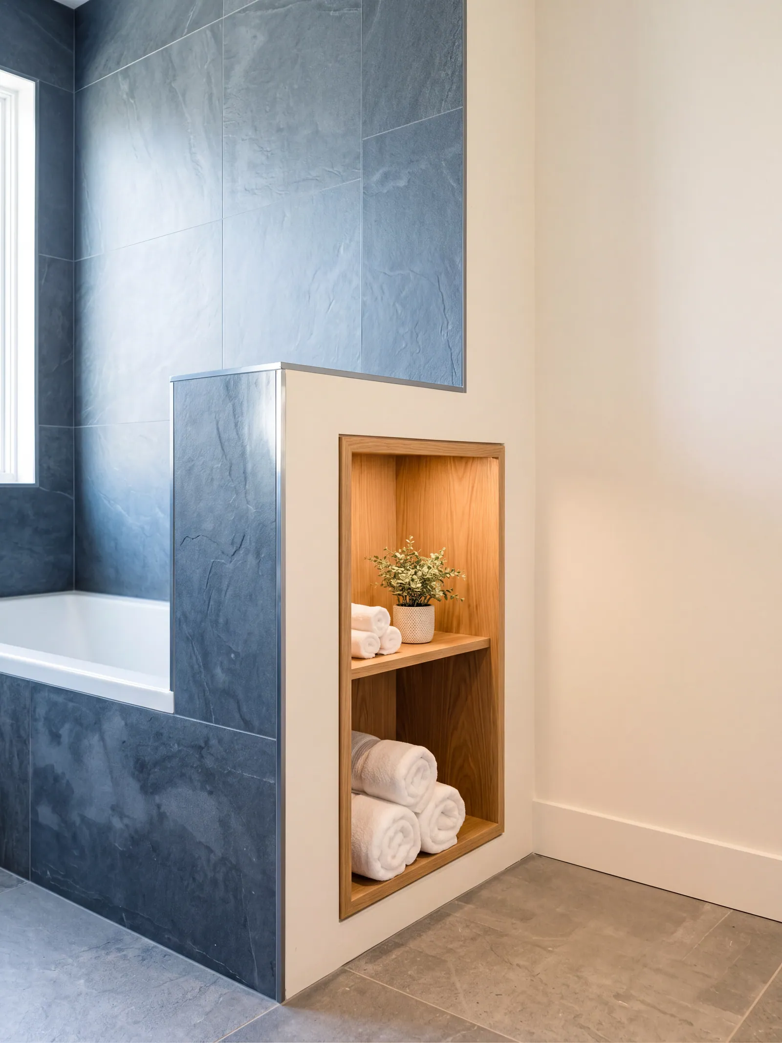 A shower wall of dark slate-look tile with a finished niche holding a wood shelf, rolled towels and a small plant, beside a tub and a window.