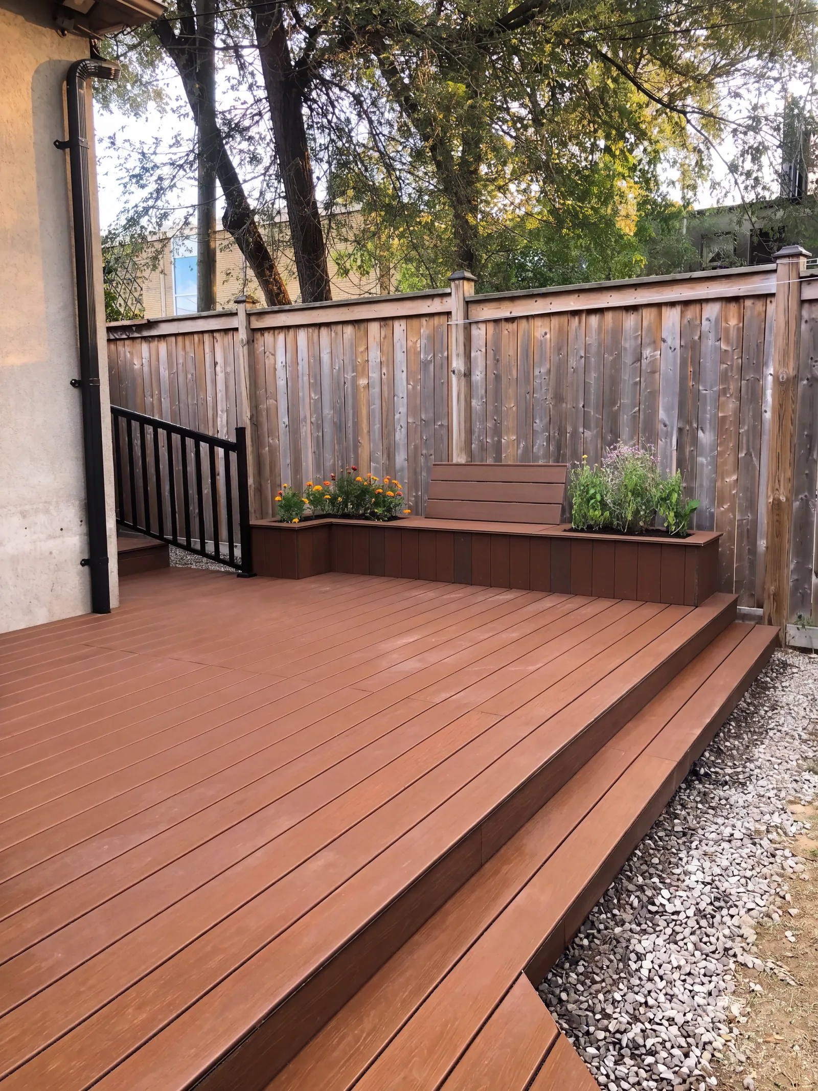 A low stained deck with a built-in planter bench along a wood fence, steps down at the corner and gravel beside it.
