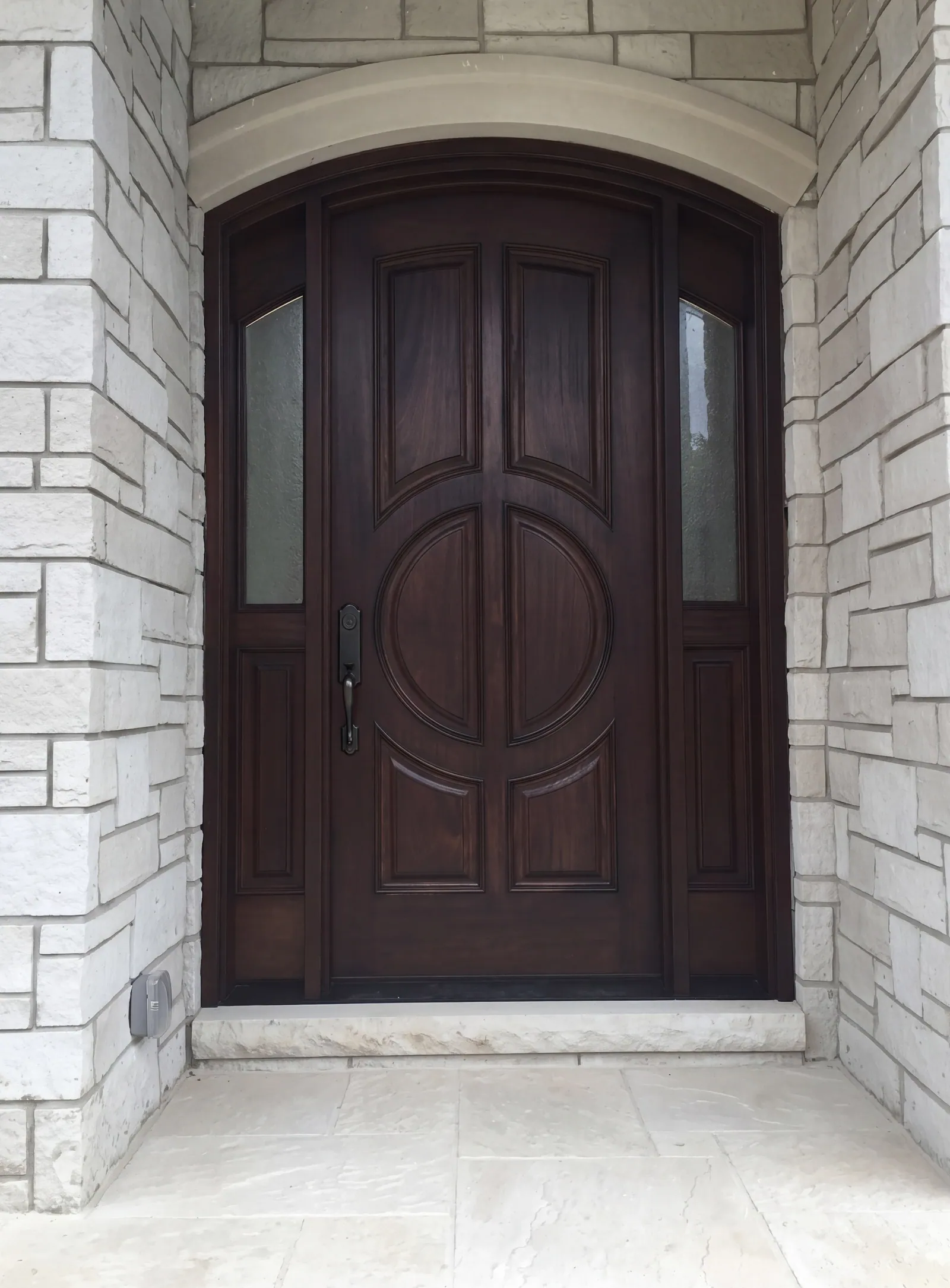 An arched dark wood front door with two glass sidelights, in a light stone surround under a curved stone arch, with a pale stone landing at the foot.