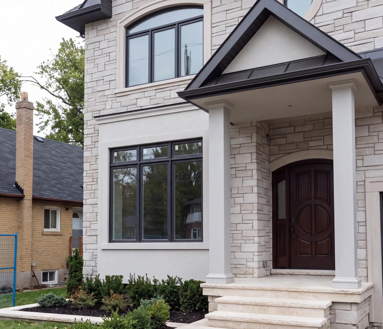 The front of a stone-clad house with an arched window, a dark-framed corner window, a gabled porch on square white columns, an arched front door and stone steps with low planting.