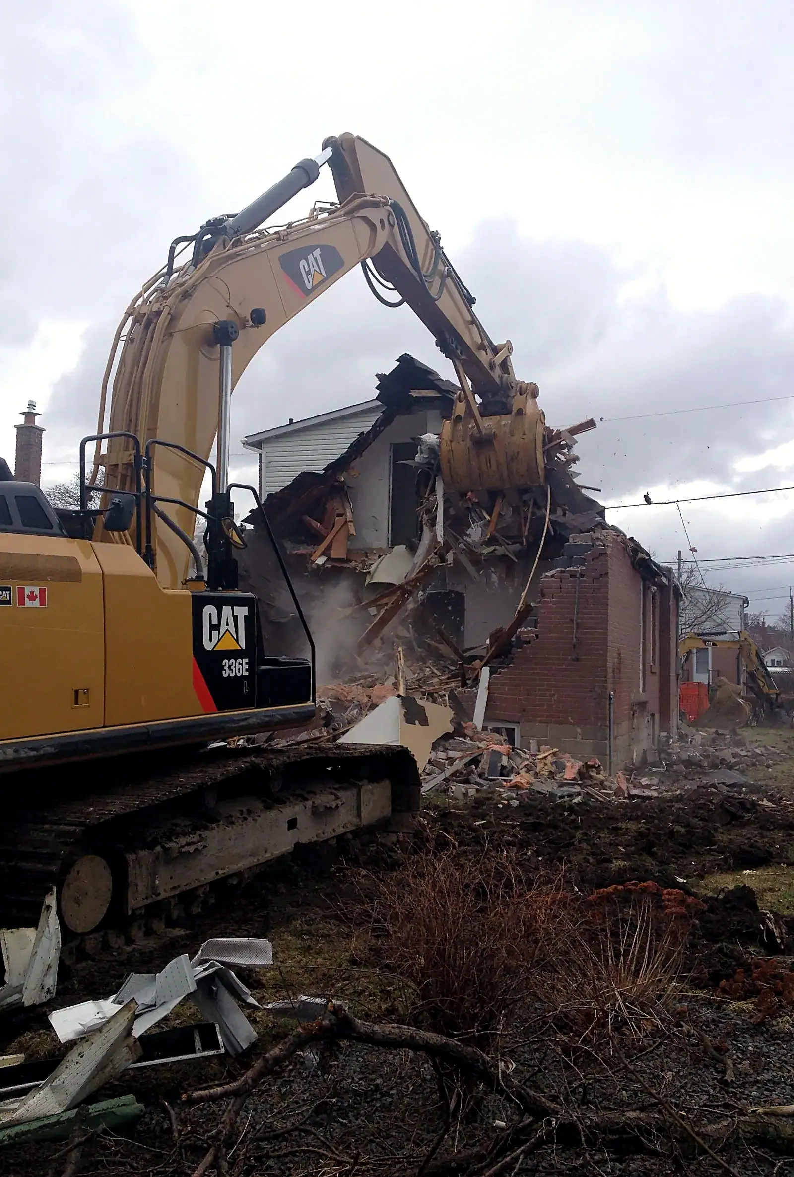 A yellow excavator with its boom raised, pulling down an old brick house, with dust and broken timber in the air.