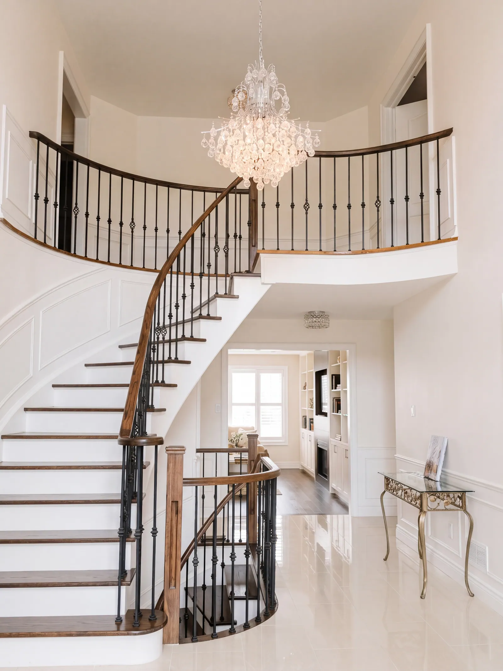 A curved stair with white risers, dark wood treads, iron balusters and a wood handrail, in a two-storey foyer under a crystal chandelier, over a polished stone floor.