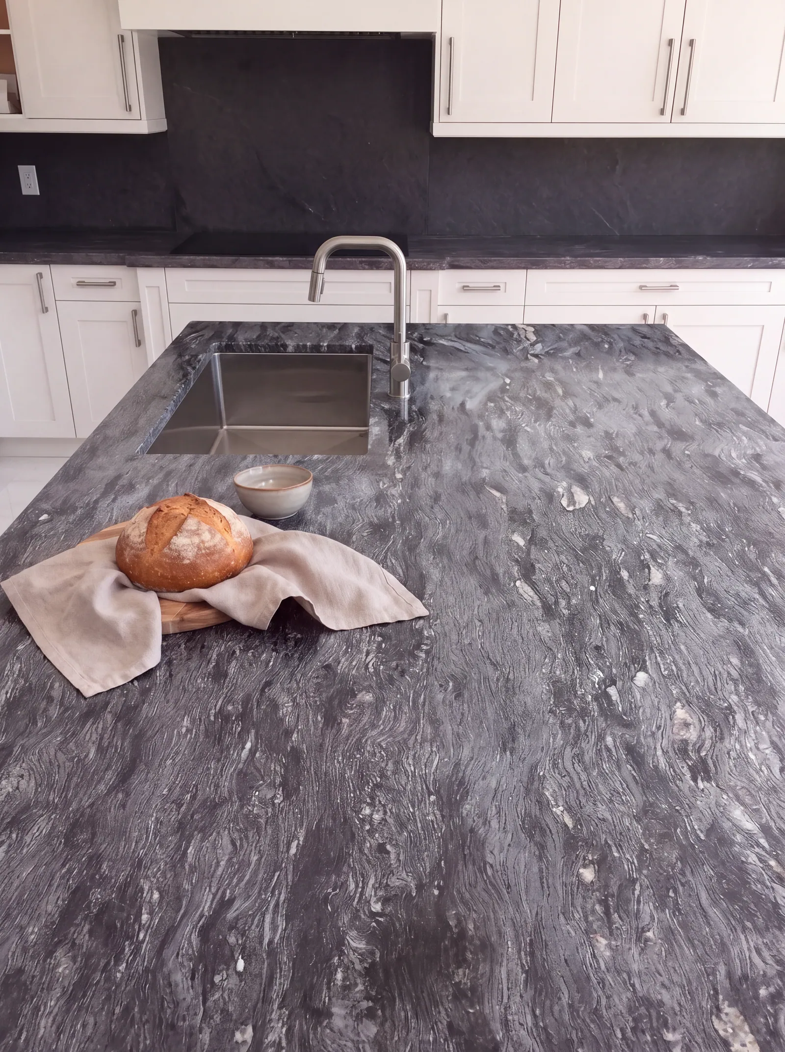 A kitchen island with a dark veined stone top that holds an undermount sink with a brushed steel faucet, a loaf of bread on a board with a linen cloth and a small bowl, in front of white cabinets and a dark stone backsplash.