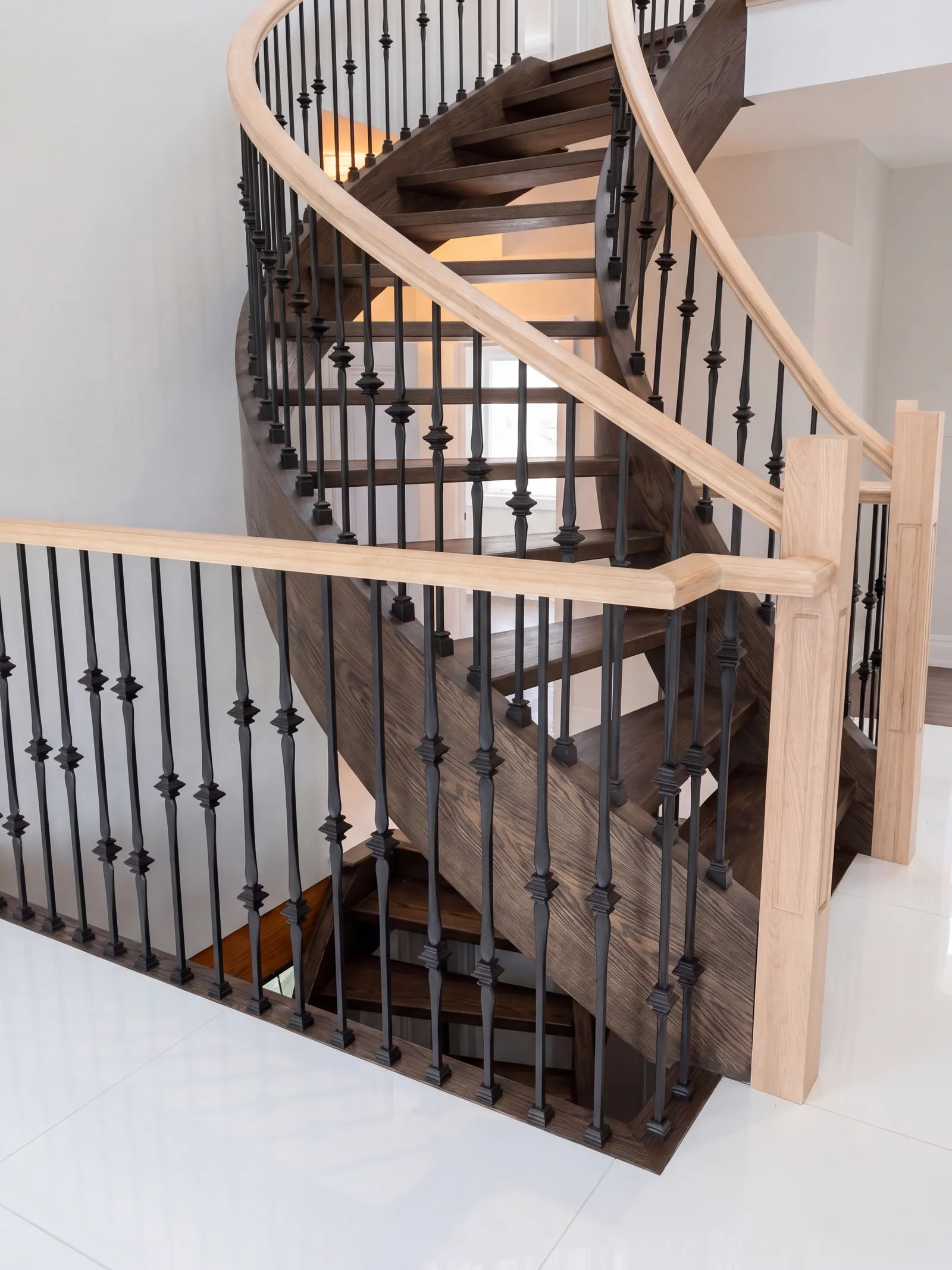 A curved stair with open dark wood treads, black iron balusters and a pale wood handrail, seen from above beside a pale wood guard rail, over a white floor.