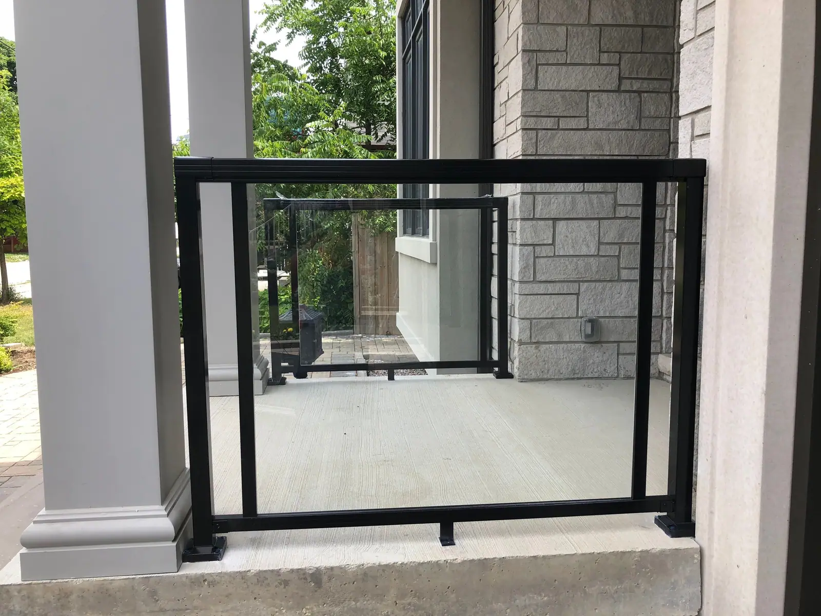 A black metal and glass railing between pale columns on a concrete porch in front of a stone-clad wall.