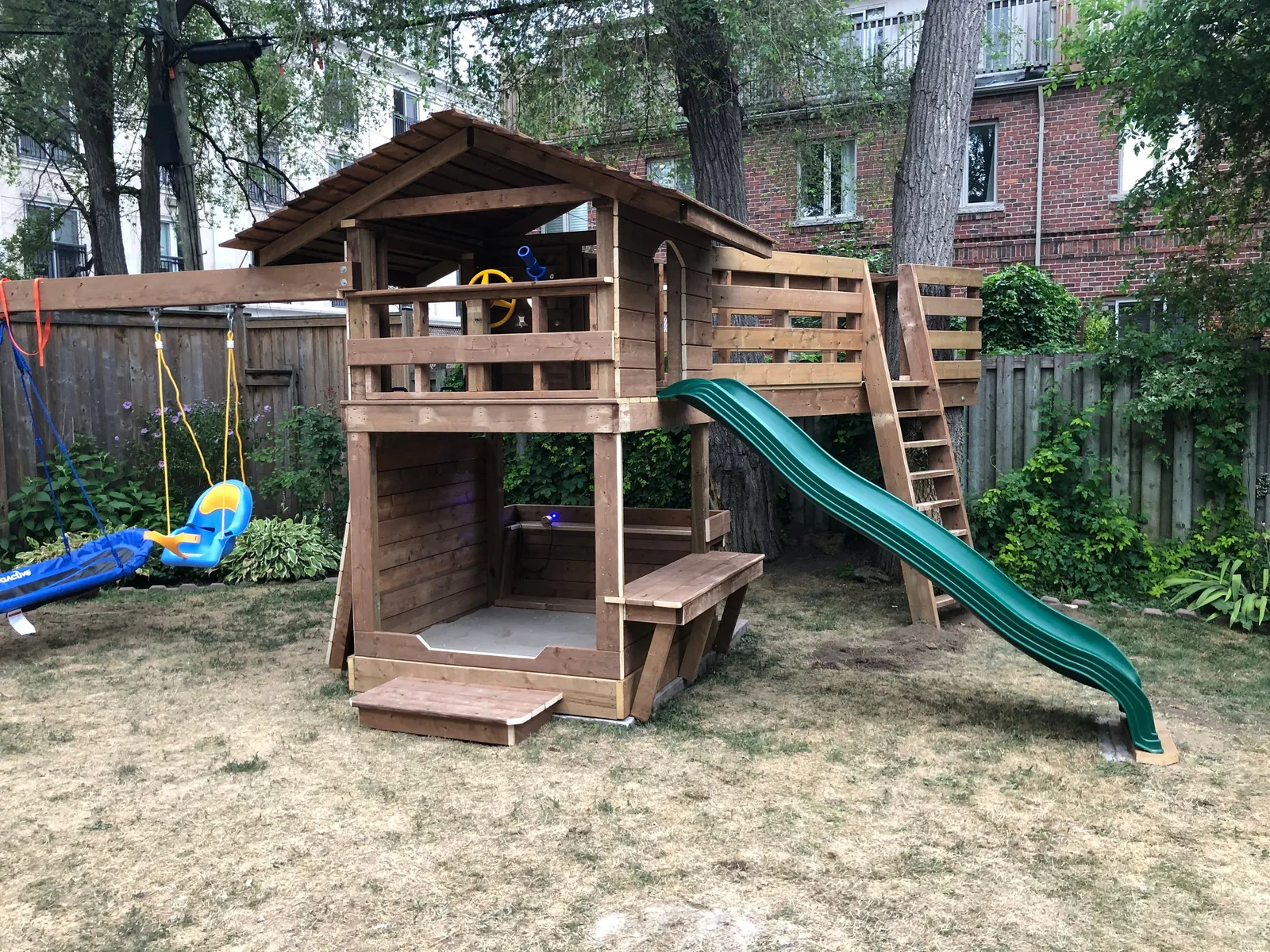 A wooden play fort with a platform, a green slide and a swing, on sandy ground.
