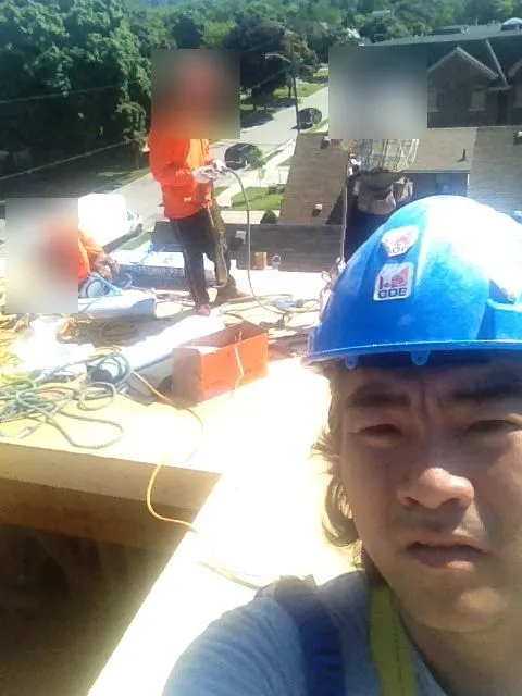 Young in a blue GDE hard hat, taking a selfie on a job site, with workers behind him on the roof.